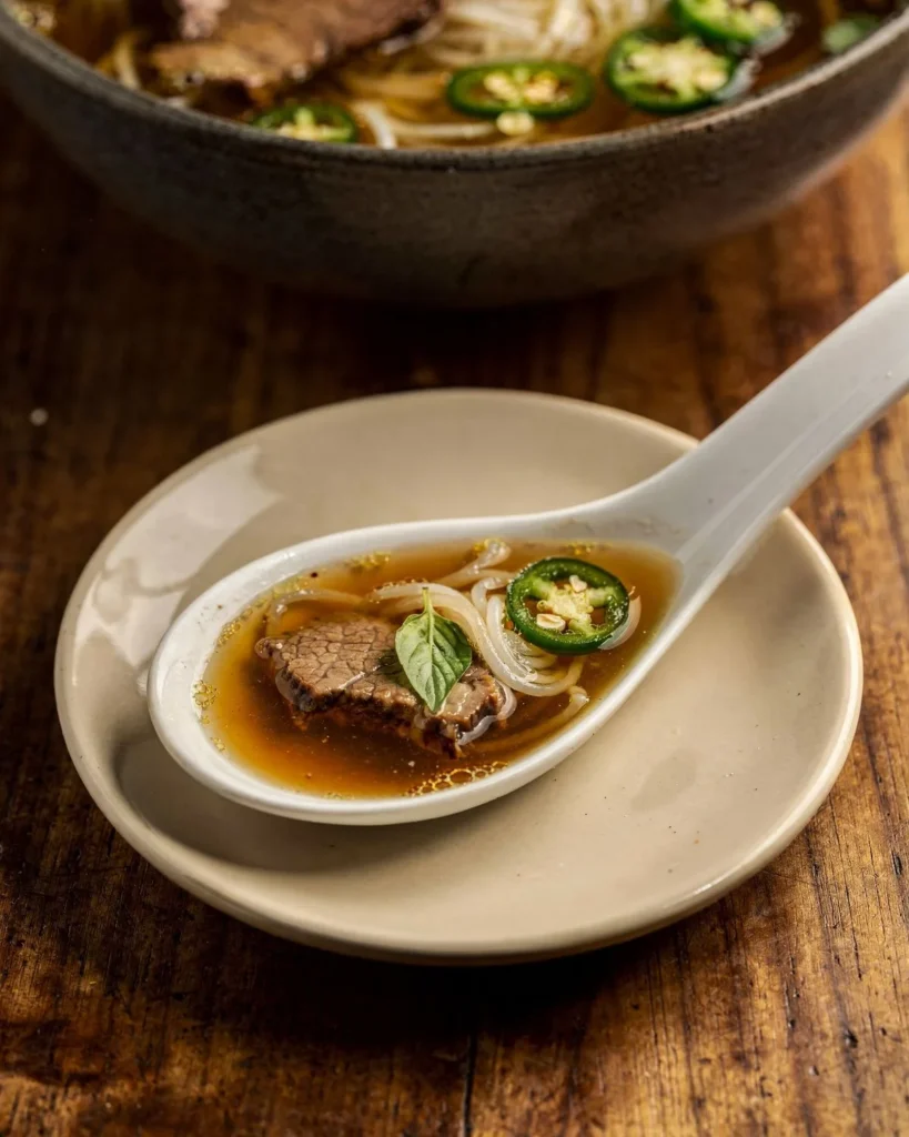 Spoonful of pho broth with tender beef, shirataki noodles, basil and jalapeno on a saucer